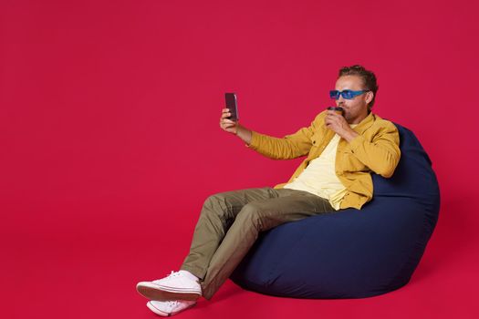 Happy handsome young man 30s make selfie using digital tablet drinking coffee from paper cup wearing casual denim outfit sitting on bag chair isolated on red background. Coffee shop business concept.