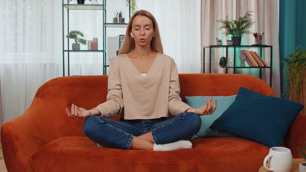 Keep calm down, relax, inner balance. Adult girl breathes deeply with mudra gesture, eyes closed meditating with concentrated thoughts, peaceful mind. Young woman sit at home in living room on couch
