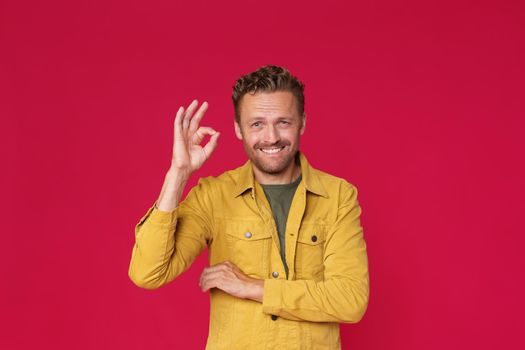 Cheerful young man showing, gesturing OK, okay smiling looking at camera wearing casual jeans yellow jacket isolated on red background. Happy handsome caucasian man in casual.