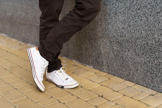 No face visible. Employee man standing outside with feet crossed wearing white running shoes take a break leaned on a wall enjoying fresh air after long hard day. Freelancer man at coffee brake.