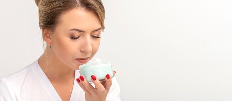 Portrait of young beautician sniffing moisturizing cream holding white jar on white background, copy space