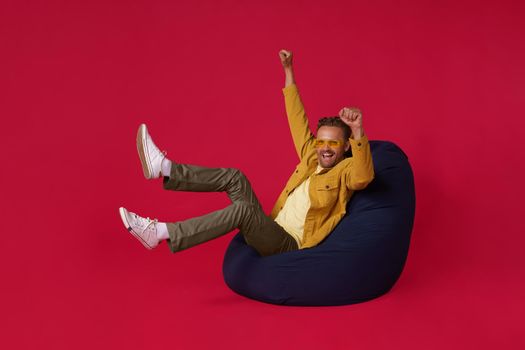 Happy handsome man sits cozy excited in victory of his sports team or business success in beanbag, bag chair wearing trendy denim outfit isolated on red background. Freelancer man success.