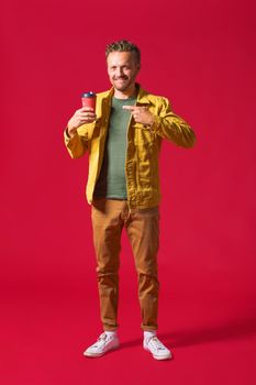 Young handsome man in casual denim clothes and white running shoes posing with paper cup of fresh coffee or tea to go isolated on red background. Happy freelancer man takeaway coffee break.