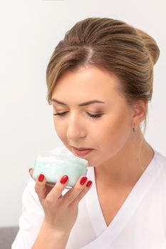 Portrait of young beautician sniffing moisturizing cream holding white jar on white background, copy space