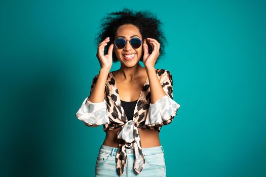 Pretty african woman with afro hair in leopard top and sunglasses smiling to camera over blue wall background. Cute mixed race girl's portrait. High quality photo