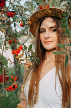 A beautiful blonde girl in a hat with long hair, on a walk in the autumn season. Portrait of a woman at a rowan tree.