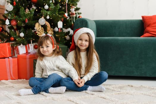 Two little cute girls sit by the Christmas tree at home and look at the camera and celebrate Christmas and New Year