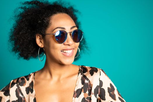 Pretty african woman with afro hair in leopard top and sunglasses smiling to camera over blue wall background. Cute mixed race girl's portrait. High quality photo