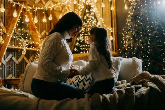 A little girl with her mother in a cozy home environment on the sofa next to the Christmas tree. The theme of New Year holidays and festive interior with garlands and light bulbs