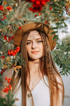 A beautiful blonde girl in a hat with long hair, on a walk in the autumn season. Portrait of a woman at a rowan tree.
