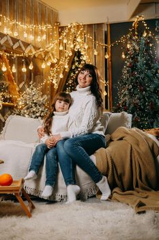 A little girl with her mother in a cozy home environment on the sofa next to the Christmas tree. The theme of New Year holidays and festive interior with garlands and light bulbs