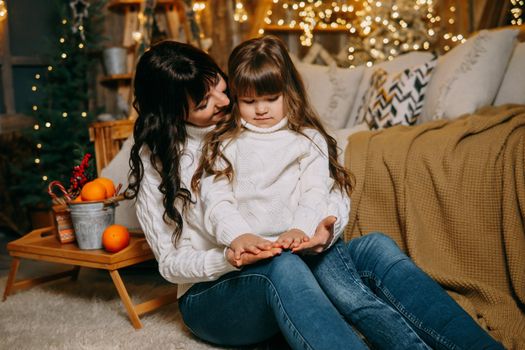 A little girl with her mother in a cozy home environment on the sofa next to the Christmas tree. The theme of New Year holidays and festive interior with garlands and light bulbs