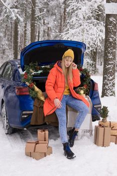A woman in a winter snow-covered forest in the trunk of a blue car decorated with Christmas decor. The concept of Christmas and winter holidays.