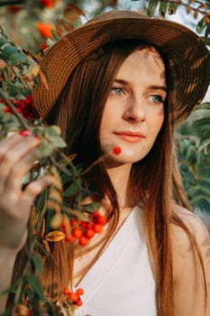A beautiful blonde girl in a hat with long hair, on a walk in the autumn season. Portrait of a woman at a rowan tree.