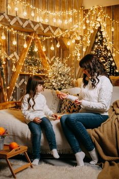 A little girl with her mother in a cozy home environment on the sofa next to the Christmas tree. The theme of New Year holidays and festive interior with garlands and light bulbs.