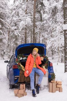 A woman in a winter snow-covered forest in the trunk of a blue car decorated with Christmas decor. The concept of Christmas and winter holidays.
