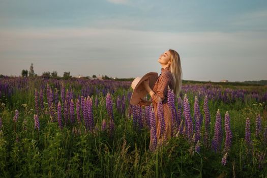 A beautiful woman in a straw hat walks in a field with purple flowers. A walk in nature in the lupin field.
