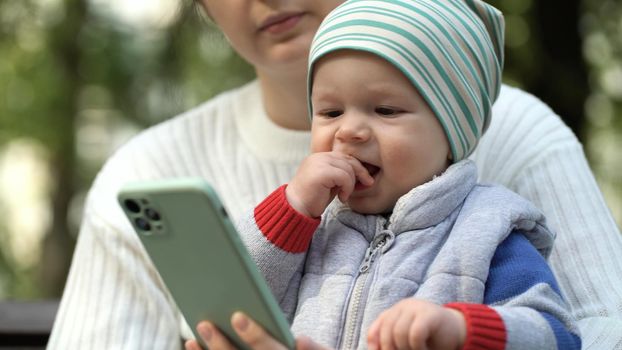 A one-year-old boy sits in his mother's arms in the park and watches a cartoon on the phone in the park close-up. Happy child waving hands while watching cartoons on smartphone.