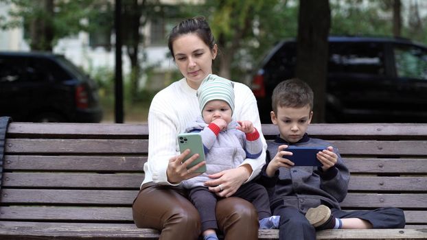 A one-year-old child sits in his mother's arms and watches cartoons in the autumn park, a seven-year-old son plays games on a smartphone.