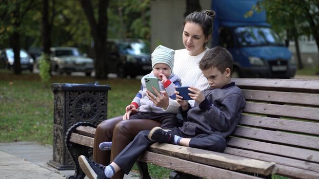 Modern family relaxing with gadgets outdoors on a bench outside. Mom and two sons in the park.