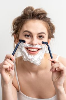 Beautiful young caucasian woman shaving her face by razor on white background. Pretty smiling woman with shaving foam and razor on her face