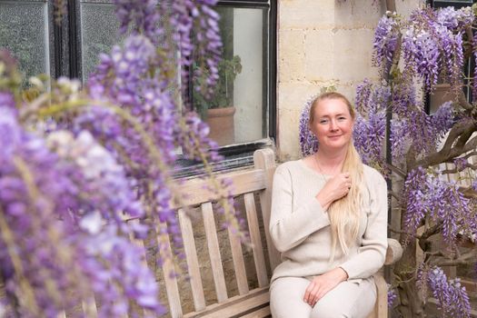 a beautiful middle-aged woman sits on a bench in the thickets of blooming wisteria and meditates happy. High quality photo