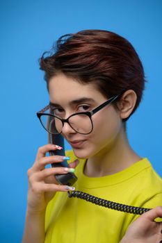 Smiling young pretty brunette guirl with extravagant nail art having conversation using old fashioned phone, studio shot at blue background