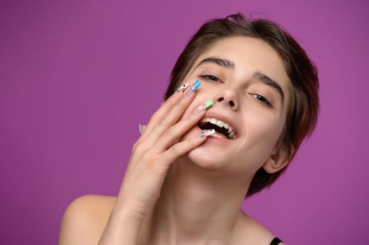 Portrait of loughing young pretty girl with right palm on her face, short haircut and extravagant nail art, magenta colored background