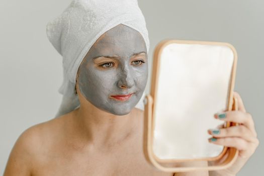 Young woman with gray clay cosmetic mask on her face looks in mirror, her hair and body wrapped in towel. Close-up