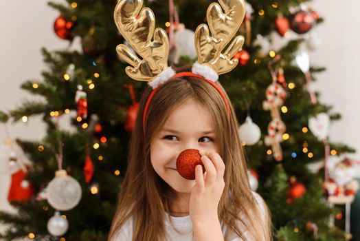 A little girl in the antlers of a Christmas deer holds a ball to her nose. Happy new year and christmas