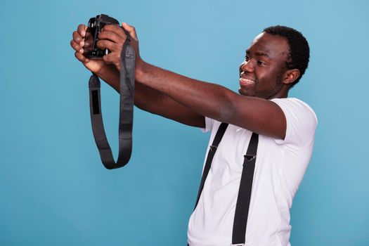 Joyful photography enthusiast taking selfie photo with DSLR camera while standing on blue background. Smiling heartily professional photographer taking picture of himself. Studio shot.