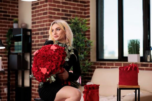 Woman posing with luxury red roses bouquet, sitting on bar stool in living room, medium shot. Beautiful blonde girl with valentines day expensive gifts, romantic date, anniversary