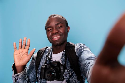 Smiling young photography enthusiast having DSLR device and taking selfie while waving at camera on blue background. Confident photographer with profesional photo device and backpack.