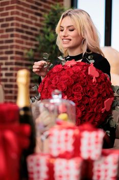 Excited woman with red roses bouquet, blurred gifts on table, valentines day luxury presents, romantic date, anniversary greeting. Smiling blonde girl sitting with flowers decor.