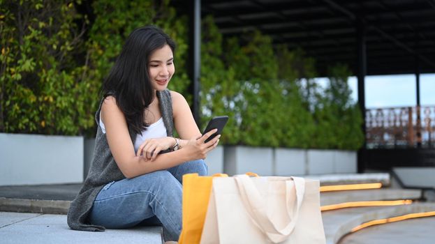 Beautiful asian woman with shopping bags using mobile phone while sitting on stairs in front of the building.