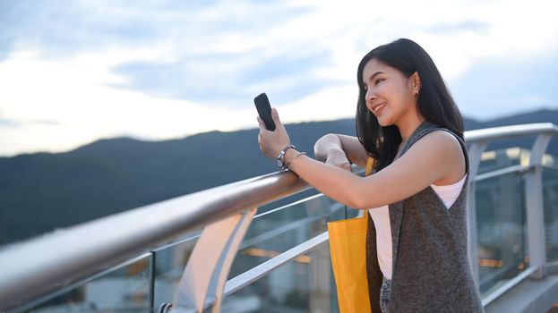 Smiling young woman typing text message on mobile phone while standing on roof terrace outdoors with cityscape on background.