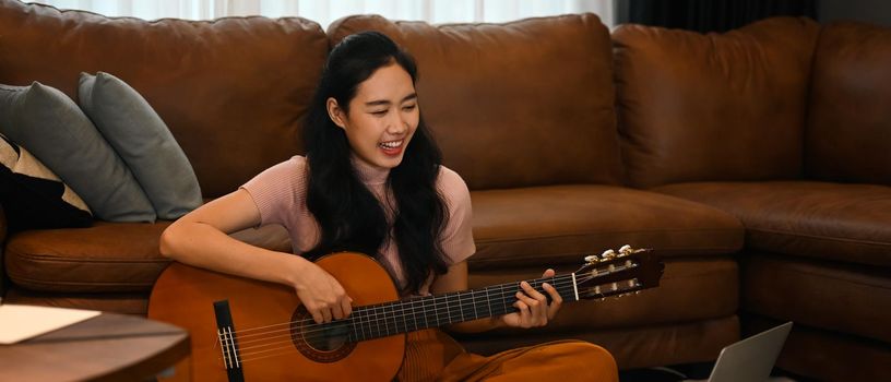 Happy young woman learning play guitar, watching online lessons on laptop. Hobbies and learning concept.