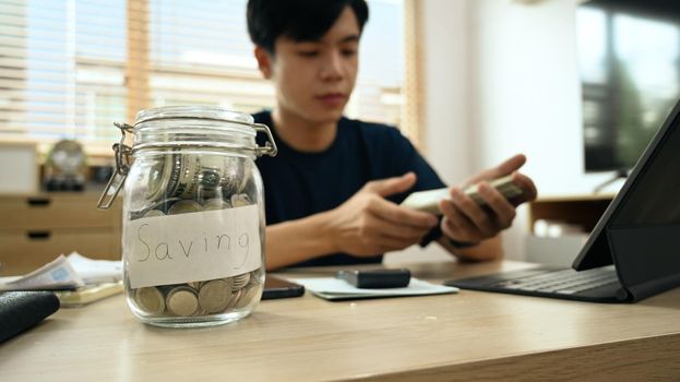 Cropped view of glass jar with coins on wooden table and man using laptop in background. Saving money and investing concept.