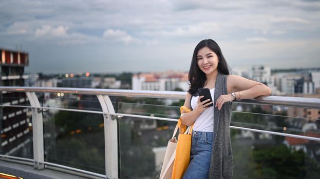 Attractive asian woman holding mobile phone standing on roof terrace outdoors with cityscape on background.