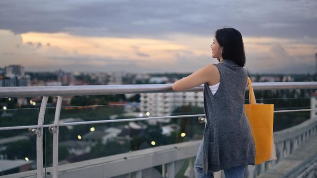 Rear view of asian woman standing on rooftop terrace with overlooking the city at sunset view.