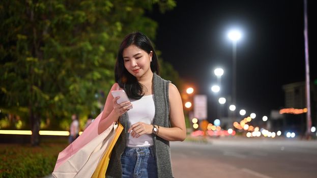 Stylish woman typing text message on her mobile phone while walking through night city street with blurred lights background.