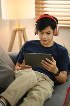Relaxed asian man in headphone using digital tablet, chatting in social networks, sitting on couch in living room.