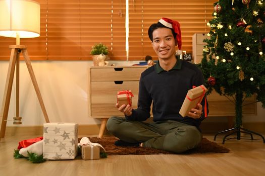 Cheerful man sitting on floor in living room and preparing Christmas gifts. Celebration, holidays and Christmas concept.