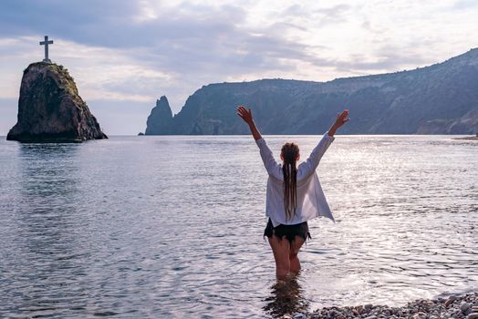 The girl stands on the shore and looks at the sea. Her hands are raised up. She wears a white shirt and her hair is in a braid
