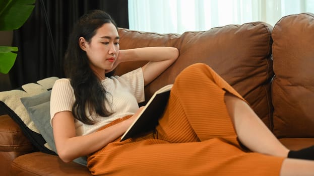 Calm asian woman reading book, relaxing in couch at home. Leisure activity, positive mood concept.
