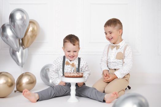 Two boys celebrating birthday, children have a B-day party. Birthday cake with candles and balloons. Happy kids, celebration, white minimalist interior