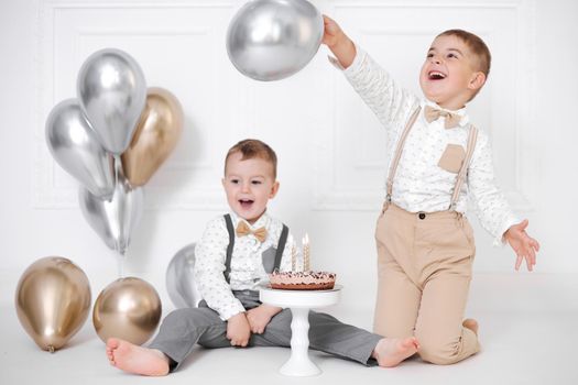 Two boys celebrating birthday, children have a B-day party. Birthday cake with candles and balloons. Happy kids, celebration, white minimalist interior
