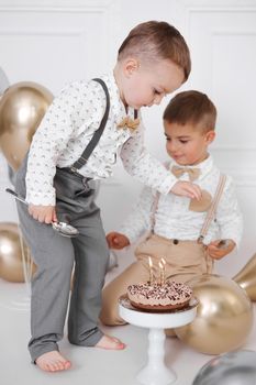 Two boys celebrating birthday, children have a B-day party. Birthday cake with candles and balloons. Happy kids eating cake, celebration, white minimalist interior