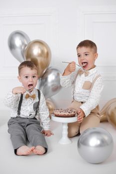 Two boys celebrating birthday, children have a B-day party. Birthday cake with candles and balloons. Happy kids eating cake, celebration, white minimalist interior