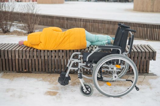 Homeless woman sleeping on a park bench next to a wheelchair